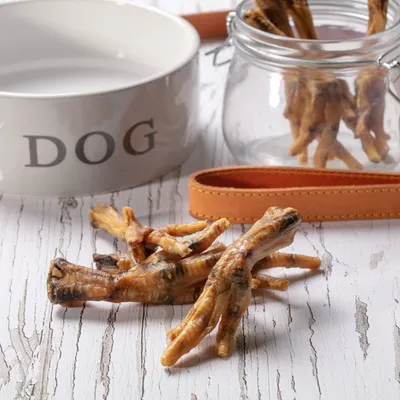 Dried poultry feet treats displayed on a wooden surface, with some in a glass jar and a bowl labelled 'DOG' nearby.