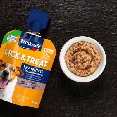 Vitakraft Lick & Treat Training Leverpølse, Made in Germany, lavt kalorieindhold, uden korn, uden tilsætning af farve- og konserveringsmidler, 60 g klempose ved siden af skål med pasta.