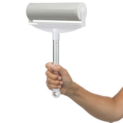 Hand houdt een grote pluizenroller met metalen handvat en witte kunststof houder, geschikt voor het verwijderen van haren of pluisjes. Roller is cilindervormig en lichtgrijs van kleur.