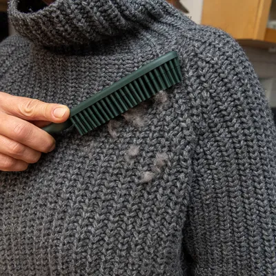 Ruka používa zelenú kefu na odstránenie zvieracích chlpov zo sivého vlneného svetra.
