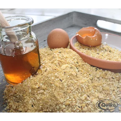 Mélange de miettes jaunes avec pot de miel, œuf entier, coquille d’œuf cassée sur assiette en terre cuite et logo Quiko visible dans le coin inférieur droit.