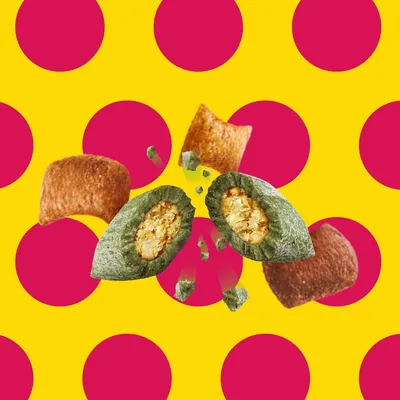 Croquettes de différentes formes et couleurs, dont une croquette verte coupée montrant un intérieur jaune, sur fond à pois rouges et jaunes. Aucun texte ou marque visible.