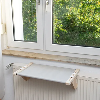 Hamac pour radiateur avec cadre en bois et tissu gris, fixé sur un radiateur sous une fenêtre.