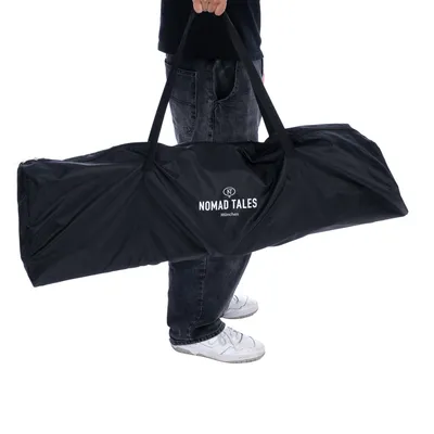 Sac de transport noir avec poignées, marque visible : NOMAD TALES München, porté à la main par une personne en jean et baskets blanches.