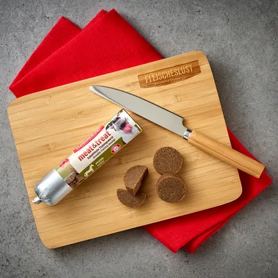 Fleischeslust meat&treat snack per cani, fette di salsiccia tagliate su tagliere di legno con coltello. Prodotto naturale, gusto cavallo.