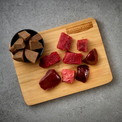 Morceaux de viande crue et abats rouges sur planche en bois marquée FLEISCHESLUST, à côté d’un bol contenant des cubes bruns. Marque FLEISCHESLUST visible sur la planche.