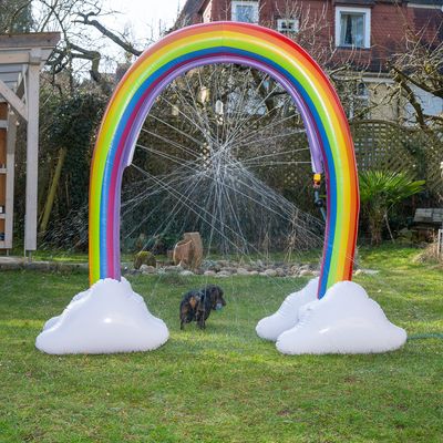 Opblaasbare regenboog-watersproeier met witte wolken, waterstralen in alle richtingen. Hond in de tuin loopt door de sproeier.