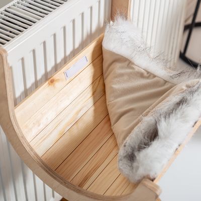 Dřevěné lehátko s měkkým béžovým polstrováním a okrajem z umělé kožešiny, připevněné k radiátoru.