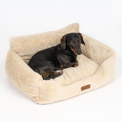 Cane sdraiato in un letto per animali beige, peloso, di forma rettangolare con lati rialzati. Etichetta in pelle con la scritta 'Designed by Lotte' visibile.