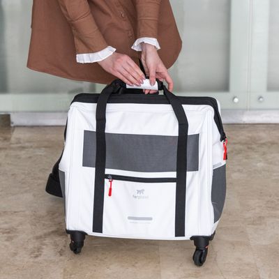 Bolsa de transporte con ruedas, blanca y negra, marca Ferplast. Mujer sostiene el asa de la bolsa. Zonas de malla y cremalleras visibles en rojo.