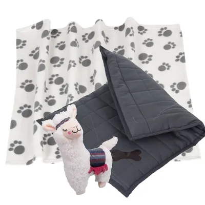 Weiße Decke mit grauen Pfotenabdrücken, dunkelgraue gesteppte Decke mit Knochenmotiv und Plüsch-Lama-Spielzeug im Vordergrund.