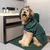 Dog wearing green bathrobe with Kooa logo, sitting on green rug in bathroom next to sink with Kooa care products.