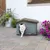 Weisse Katze verlässt ein graues Kunststoff-Katzenhaus mit braunem Dach, aufgestellt auf einer Terrasse mit Blumen im Hintergrund.