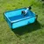 Piscina retangular azul para cães, estrutura em metal preto, com cão pequeno no interior. Produto colocado sobre relva verde.
