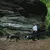 Zwei Hunde an Leinen mit Geschirren, geführt von Personen in einer natürlichen Umgebung mit Felsen und Wald.