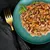 Close-up of a green plate with wet food, containing meat pieces, carrots and peas, garnished with basil. Golden fork and black cloth beside it.