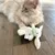 Giocattolo di peluche a forma di coniglio bianco con collare verde ed etichetta 'Müller' su pavimento in legno. Gatto sfocato sullo sfondo.