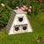 Driehoekig wit houten huis met drie kattenvormige ingangen, geschikt voor de tuin. Dak met grijze shingles, omgeven door bloemen en gras.