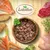 Bowl of wet pet food surrounded by visible ingredients including salmon, beef, chicken, potatoes, carrots, peas, and broccoli. Lukullus brand name shown above.