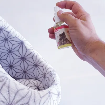 Mano sosteniendo un spray Beaphar CatComfort con imagen de gato en la etiqueta, junto a una cama acolchada blanca con patrón geométrico gris visible.