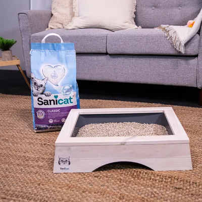 Sac de litière pour chat Sanicat Classic à côté d’un bac à litière en bois rempli, posé sur un tapis devant un canapé gris. Marque et nom du produit visibles sur le sac et le bac.