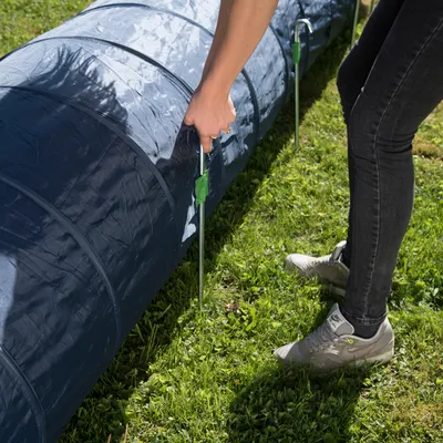 Persona fijando un túnel de tela azul al suelo con piquetas metálicas sobre césped, mostrando el montaje del producto para mascotas.