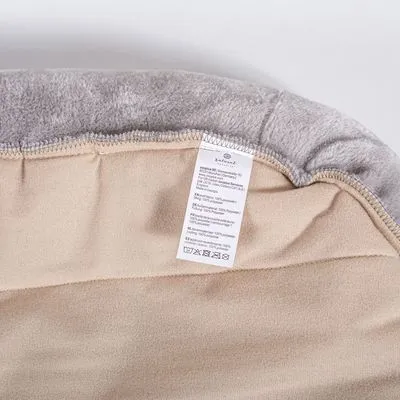 Binnenkant van een beige en grijs huisdierbed met zichtbaar waslabel, materiaal lijkt stof. Label toont onderhoudssymbolen en tekst in meerdere talen.