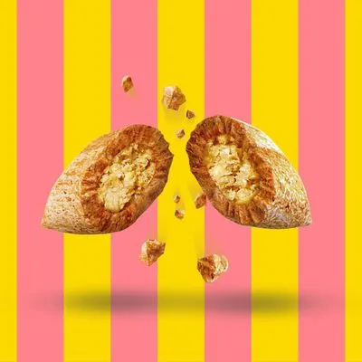 Przekrojony krokiet lub przysmak dla zwierząt z widocznym nadzieniem, kawałki rozsypane wokół. Tło w różowo-żółte pasy.