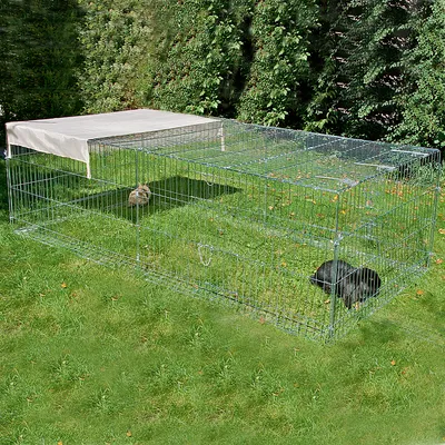 Groot metalen buitenverblijf met traliewanden en afneembare zonwering, twee konijnen in het verblijf op een groen grasveld.