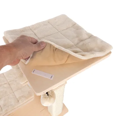 Abnehmbare, beige Plüschauflage mit Klettverschluss auf einer Holzplattform. Hand hebt die Auflage an, darunter sichtbarer Klettstreifen.