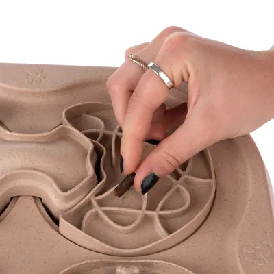 Hand placerar godisbit i beige aktivitetsleksak av plast med labyrintmönster för husdjur.