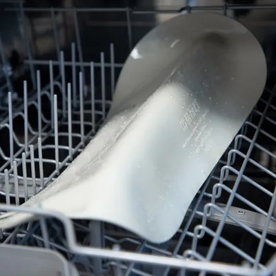 Pelle en plastique gris clair avec inscriptions visibles, posée dans le panier supérieur d’un lave-vaisselle après lavage, gouttes d’eau présentes sur la surface.