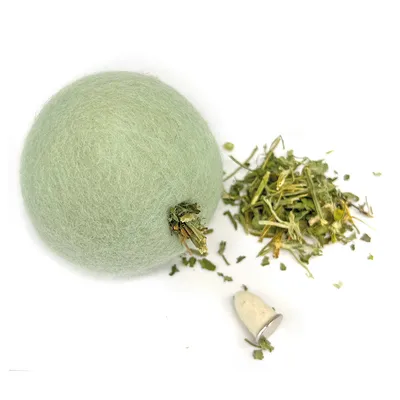 Balle en feutre verte avec herbes et insert amovible, herbes en vrac et pièce métallique visibles à côté.