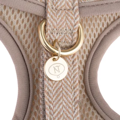 Nærbillede af en beige-gul sele med netmateriale, guldfarvet D-ring og vedhæng med NT-logo.