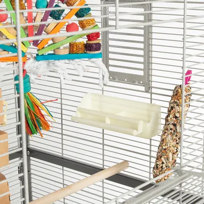 Vogelkäfig mit Spielzeug aus buntem Holz, weißem Seil und Futterhalterung. Hirsekolben als Snack befestigt. Sitzstange aus Holz sichtbar.