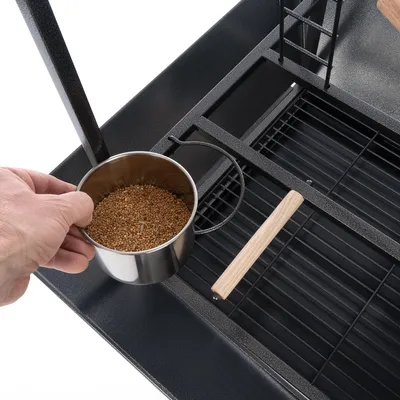 Hand hält einen Metallnapf mit Körnern über einem schwarzen Gitter, Teil eines Vogelkäfigs mit Holzstange und Metallrahmen.