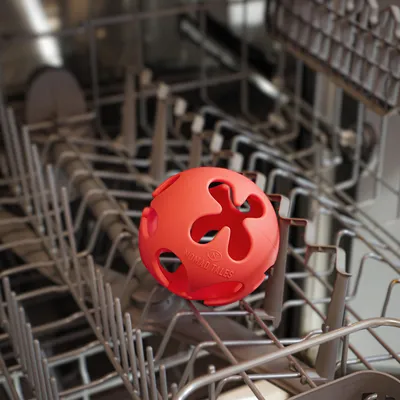 Balle rouge en plastique avec inscription 'NOMAD TALES' placée dans un lave-vaisselle ouvert, montrant la compatibilité avec le lavage en machine.