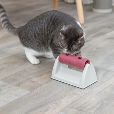 Kat bij interactief speeltje van wit plastic met rode rol en openingen, geschikt voor het verstoppen van snacks of brokjes. Product wordt door kat onderzocht op houten vloer.