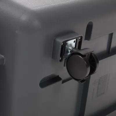 Roue noire en plastique fixée sur un angle d’une caisse ou boîte grise, fixation métallique visible. Aucun texte ou marque apparente.