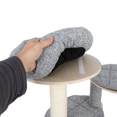 Main plaçant un coussin gris amovible sur une plateforme en bois d’un arbre à chat, fixation par bande auto-agrippante visible. Revêtement tissu, poteau recouvert de corde.