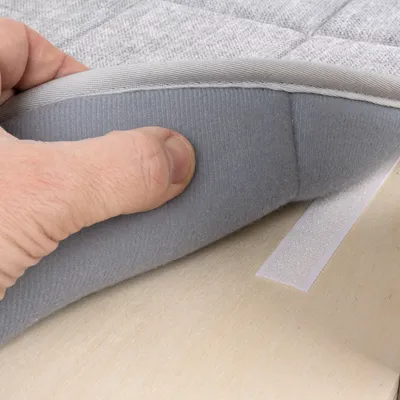 Close-up of a hand lifting a grey padded mat with a hook-and-loop fastener. Shows the mat's attachment method.