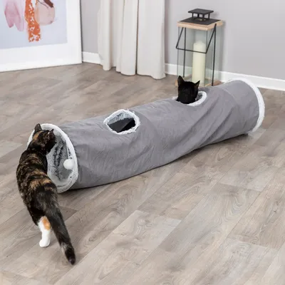 Grauer Katzentunnel mit zwei Öffnungen und Plüschrand, eine Katze spielt am Eingang, eine andere schaut aus der mittleren Öffnung heraus.