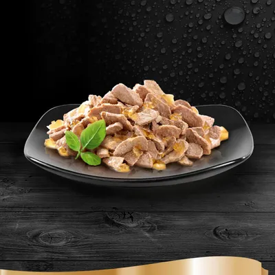 Chunks of meat in gravy served on a black plate with a garnish of green leaves, displayed against a dark background.