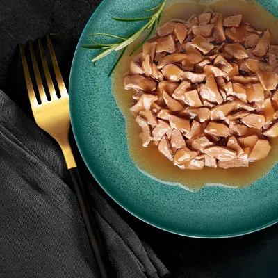 Närbild av en grön tallrik med saftiga köttbitar i sås, dekorerad med en rosmarinkvist. Guldgaffel och svart servett bredvid.