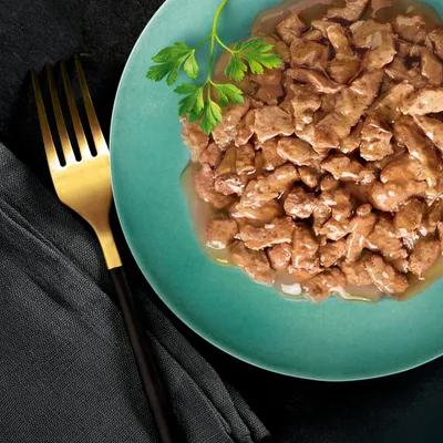 Assiette verte contenant des morceaux de nourriture humide en sauce, garnie d'une branche de persil. Fourchette dorée posée à côté sur une serviette noire.