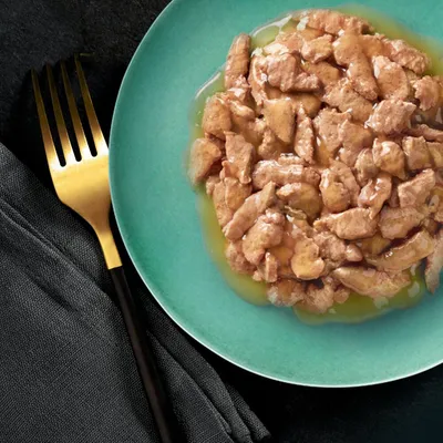 Morceaux de nourriture humide en sauce servis dans une assiette verte, fourchette dorée à côté sur une serviette noire. Aucun emballage ou marque visible.