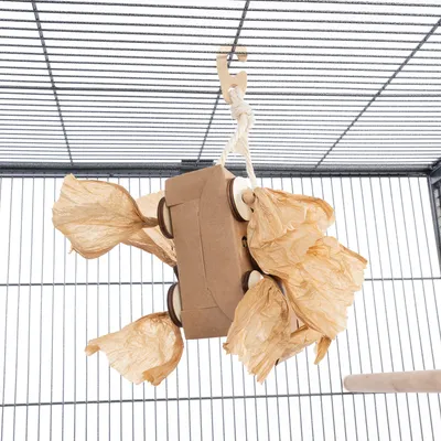 Jouet suspendu pour oiseaux en carton et papier, accroché à une cage par une corde blanche avec crochet. Plusieurs éléments froissés en papier brun visibles autour du jouet.