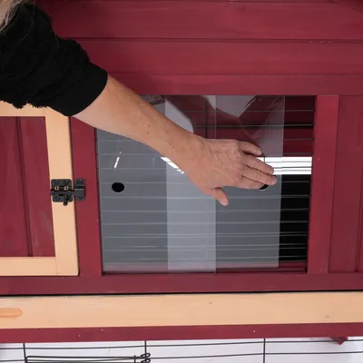 Hand zeigt Plexiglas-Schiebetür eines roten Kleintierstalls mit Holzrahmen und Metallverschluss.