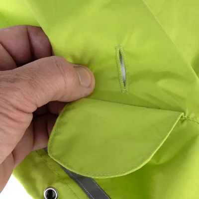 Détail d’une main tenant un vêtement vert en tissu avec fente cousue et œillet métallique visible, couture apparente. Aucun texte ou marque visible.