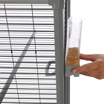 Hand attaches transparent feeder with seeds to grey metal cage. Shows easy assembly of the feeder.